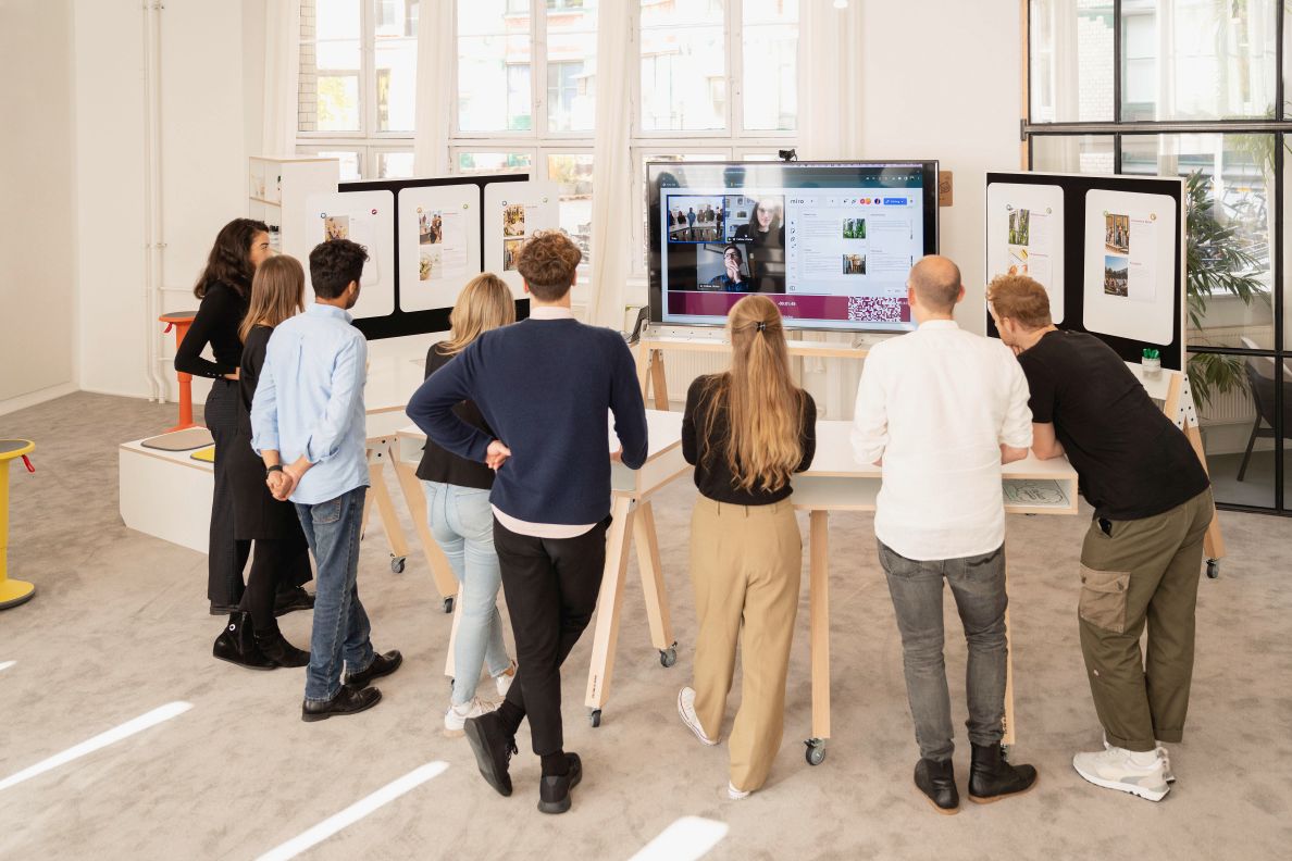 Gruppe von Personen nutzt What if We Fly Taskboard Medienwand mit Whiteboard und Bildschirm für Präsentation.