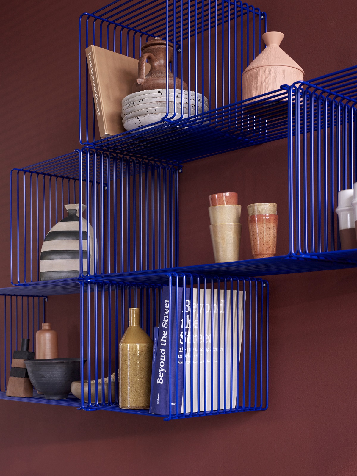 Blaues Panton Wire Regalsystem mit Deko-Objekten an einer roten Wand. Modulares Regal für Wohnraumgestaltung.