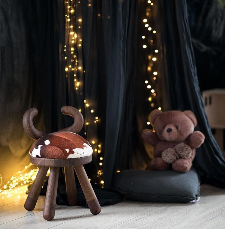 Cow Chair Kinderstuhl mit Kuhfell-Sitzfläche und Holzbeinen vor dunklem Hintergrund mit Lichterkette.