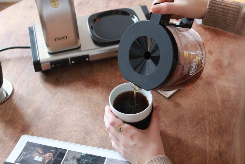 Frau gießt Kaffee aus einer Glaskaraffe in eine Tasse, Kaffeewärmer im Hintergrund.