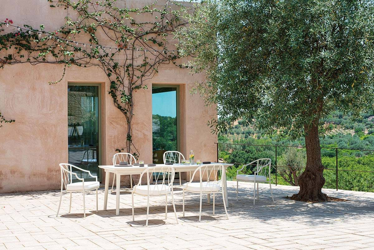 Emu Plus4 Ausziehtisch im Außenbereich mit weißen Stühlen auf einer Terrasse unter einem Baum.