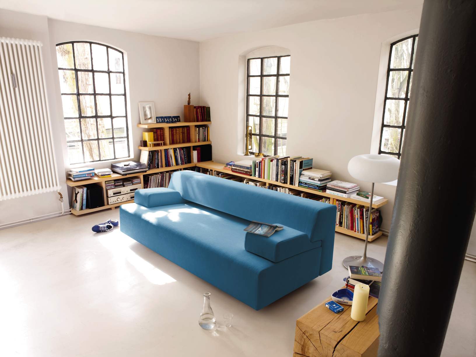 Blaues Cosma Schlafsofa in modernem Wohnzimmer mit Bücherregalen und großen Fenstern. Bequeme Couch zum Schlafen.