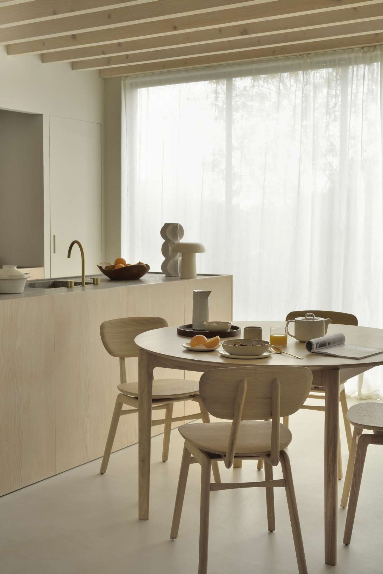 Heller Essbereich mit rundem Holztisch, Stühlen und Frühstücksgeschirr vor einem Fenster mit Vorhang.