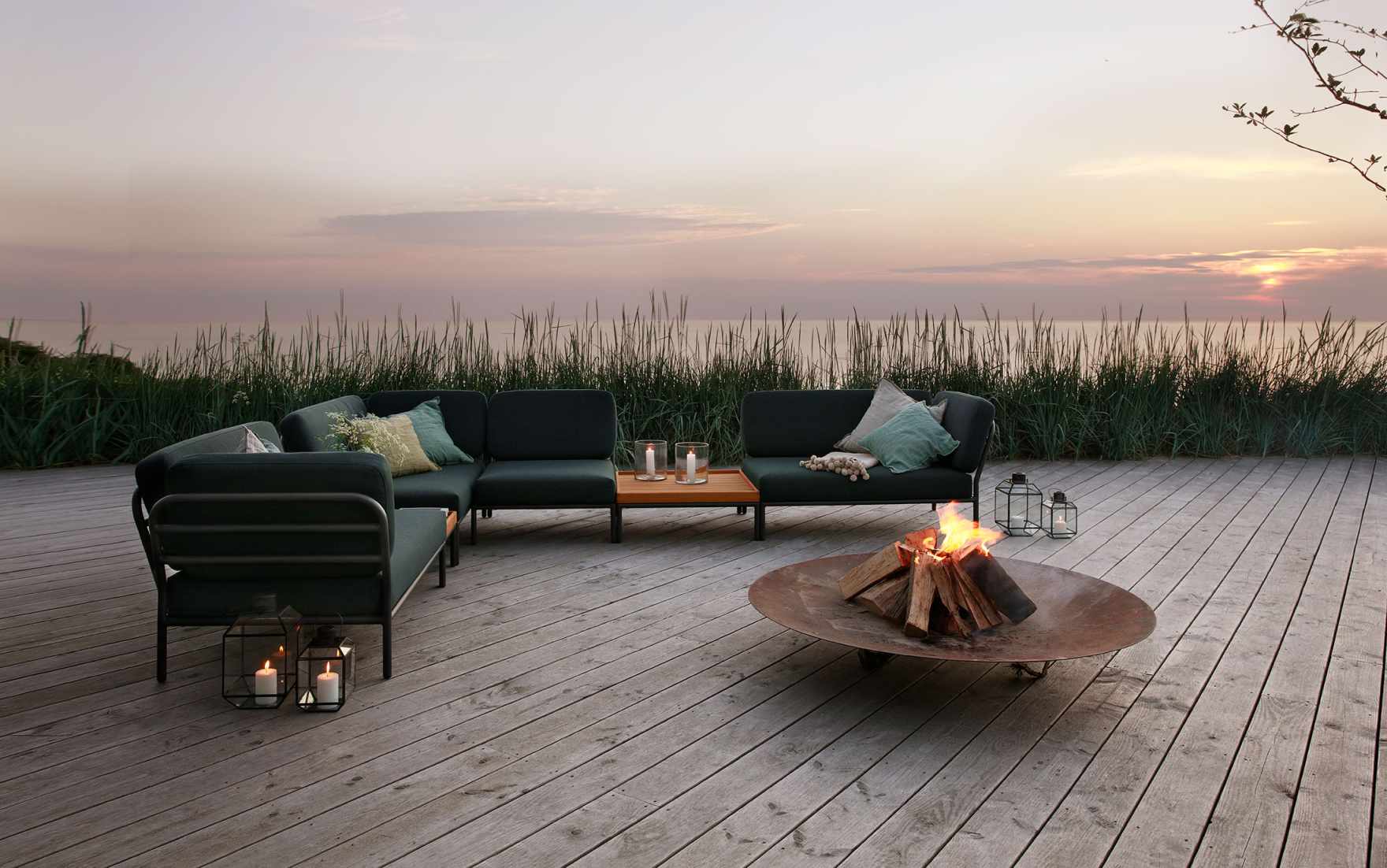 Houe Level Outdoor Sofa Ecke links auf Holzterrasse mit Feuerstelle und Meerblick.