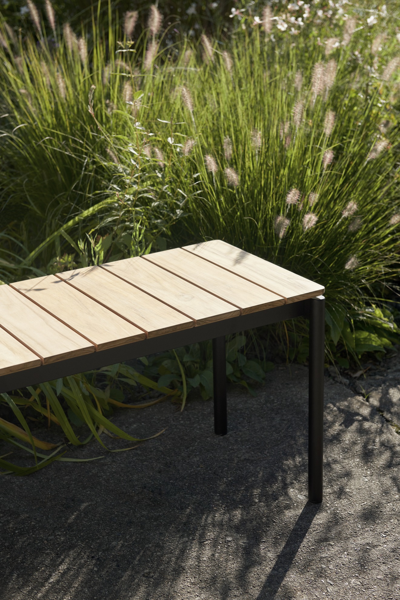 AndTradition Ville AV28 Gartenbank aus Teakholz und schwarzem Stahlgestell vor üppigem Grün.