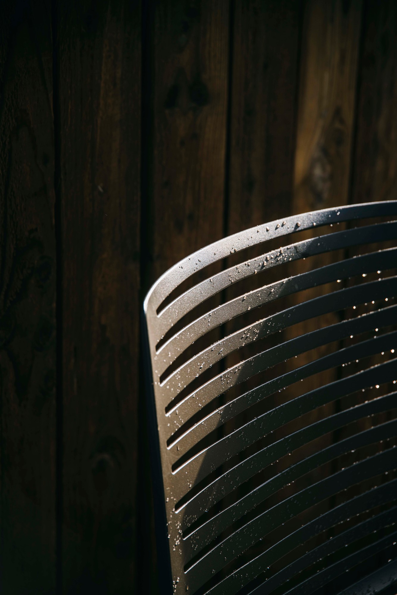 Detailaufnahme eines Zebra Outdoor Stuhls Fast aus Metall mit Wassertropfen vor dunkler Holzwand.