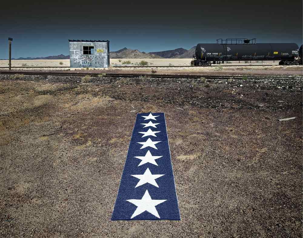 Blauer Teppich mit weißen Sternen auf einem staubigen Boden vor einer Eisenbahnstrecke und Waggons.