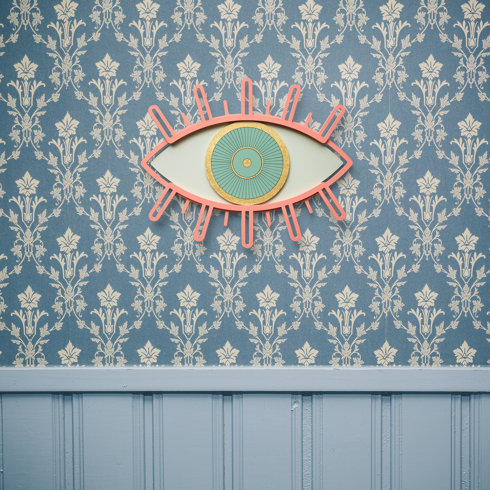 Eye 5 Wanddeko: Dekoratives Auge aus Holz in Rosa, Gold und Türkis an gemusterter Wand.