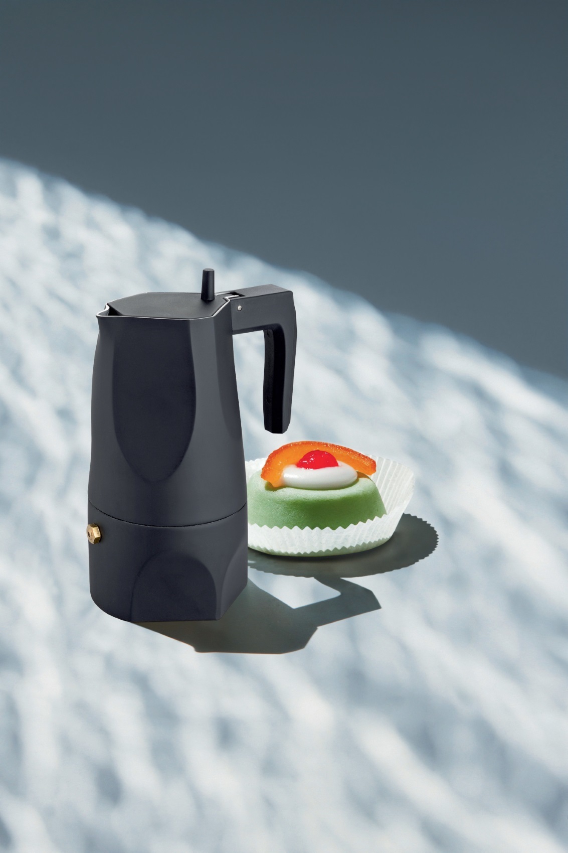 Schwarzer Alessi Espressokocher "Ossidiana" neben einem grünen Gebäckstück, ideal für die Kaffeezubereitung.