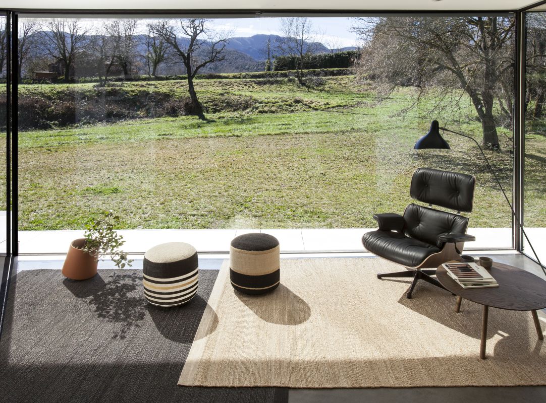 Brauner Nanimarquina Herb Teppich (250x350cm) im modernen Wohnzimmer mit Sessel und Blick ins Grüne.