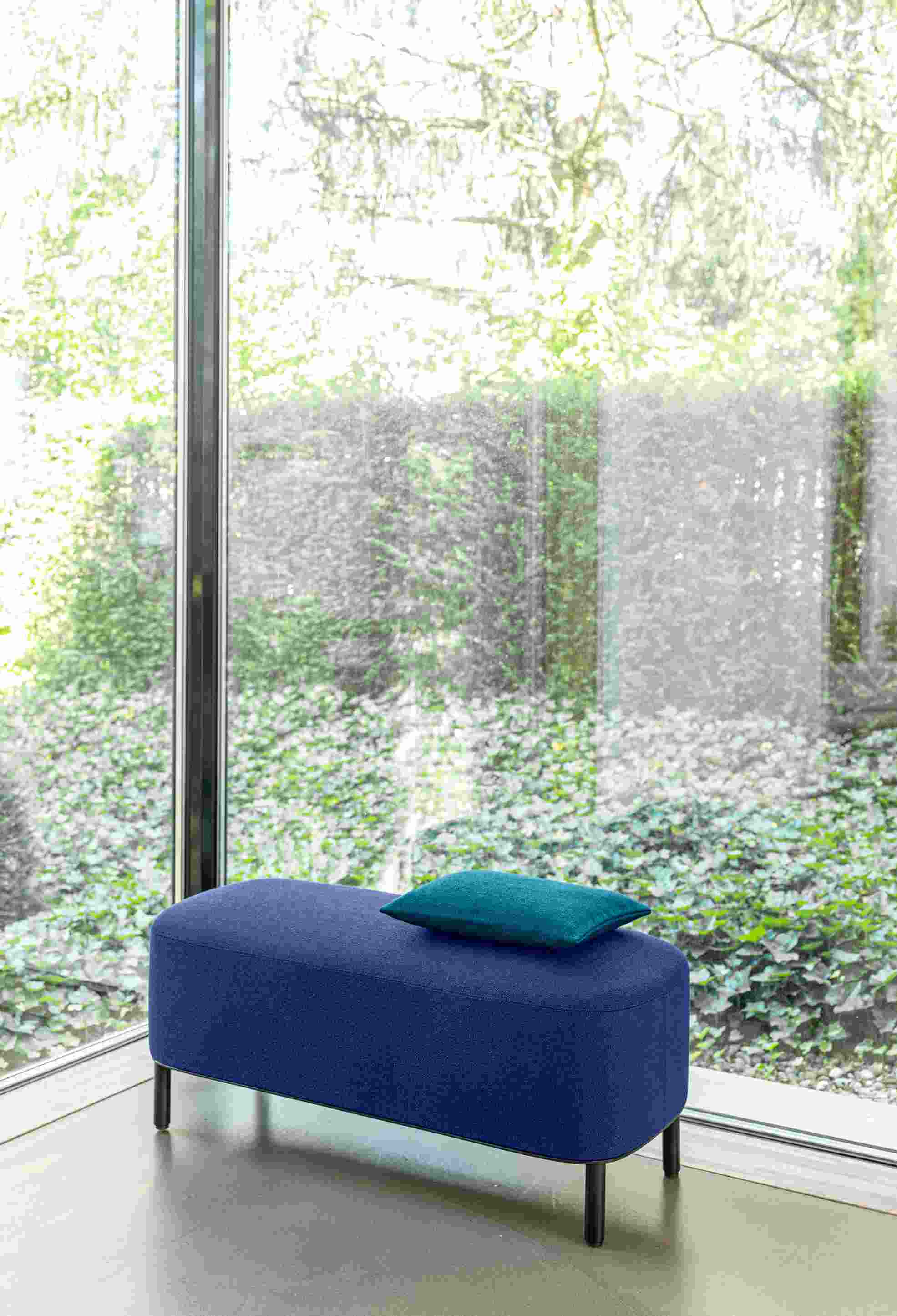Blaue Amie Bank von Schönbuch mit türkisfarbenem Kissen vor großem Fenster. Moderne Sitzbank für Wohnzimmer.
