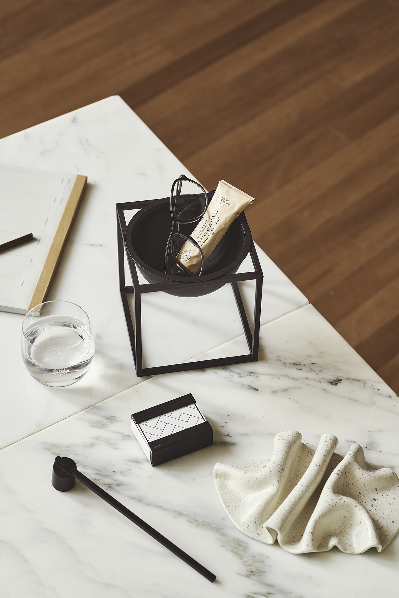 Stillleben auf Marmortisch: Handcreme, Brille in Schale, Notizbuch, Wasserglas und dekorative Keramik.