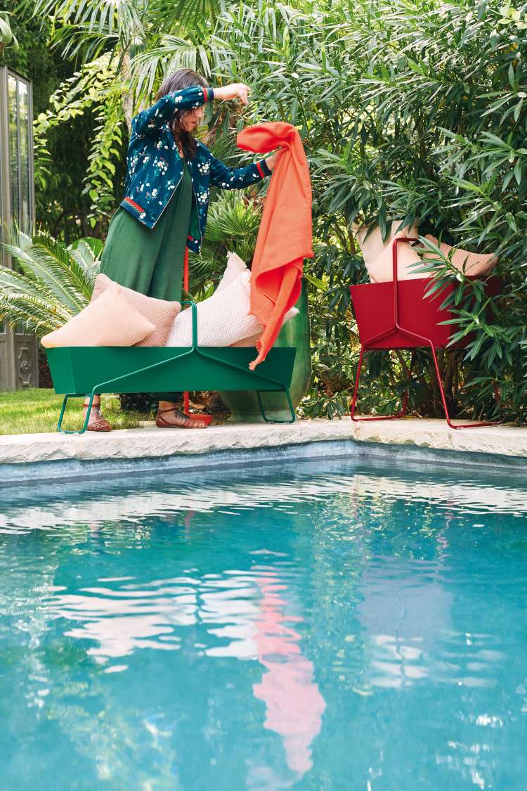 Frau drapiert Tuch über grünen Fermob Basket Blumenkasten am Pool. Roter Blumenkasten im Hintergrund.