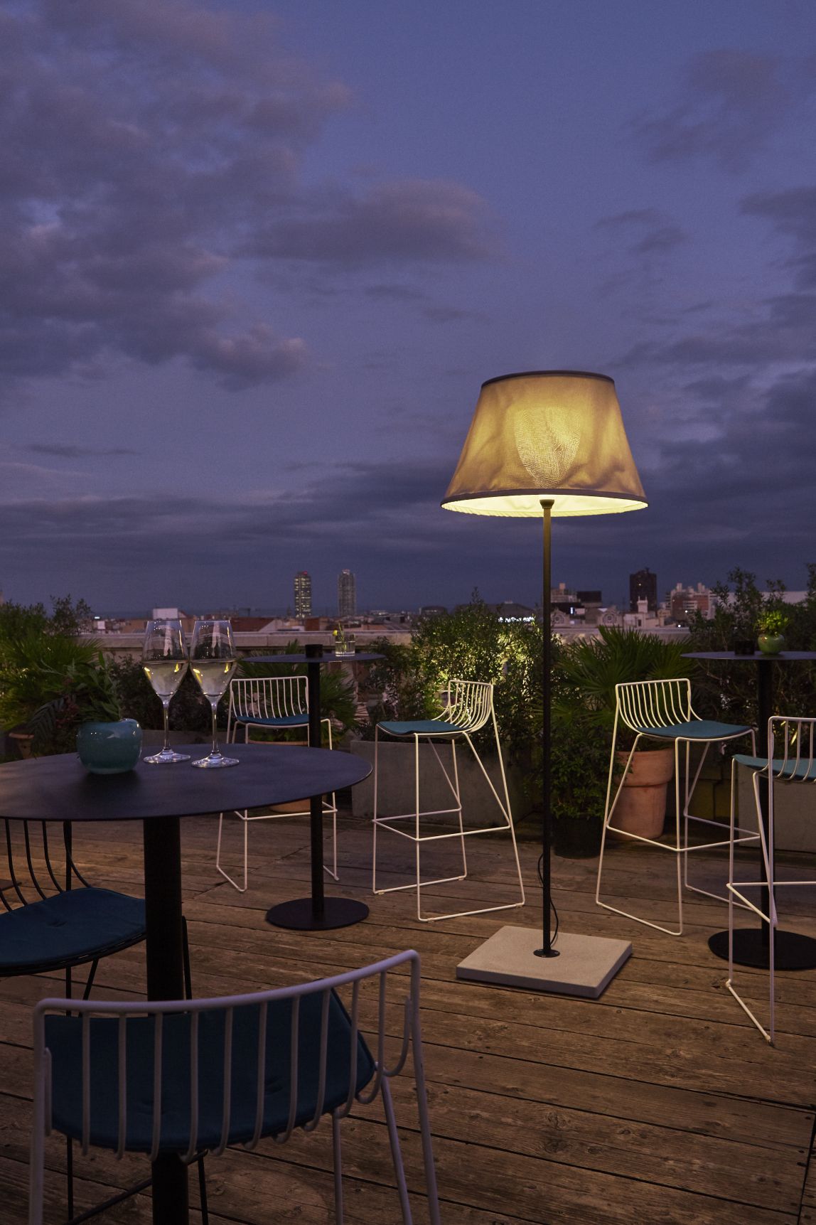 Beige TXL Stehleuchte von Marset beleuchtet abendliche Dachterrasse mit Barhockern und Skyline im Hintergrund.