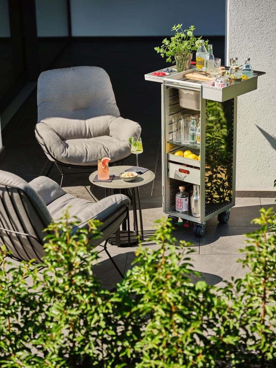 Silberner Barwagen mit Getränken und Gläsern auf einer Terrasse mit Sesseln und Beistelltisch.