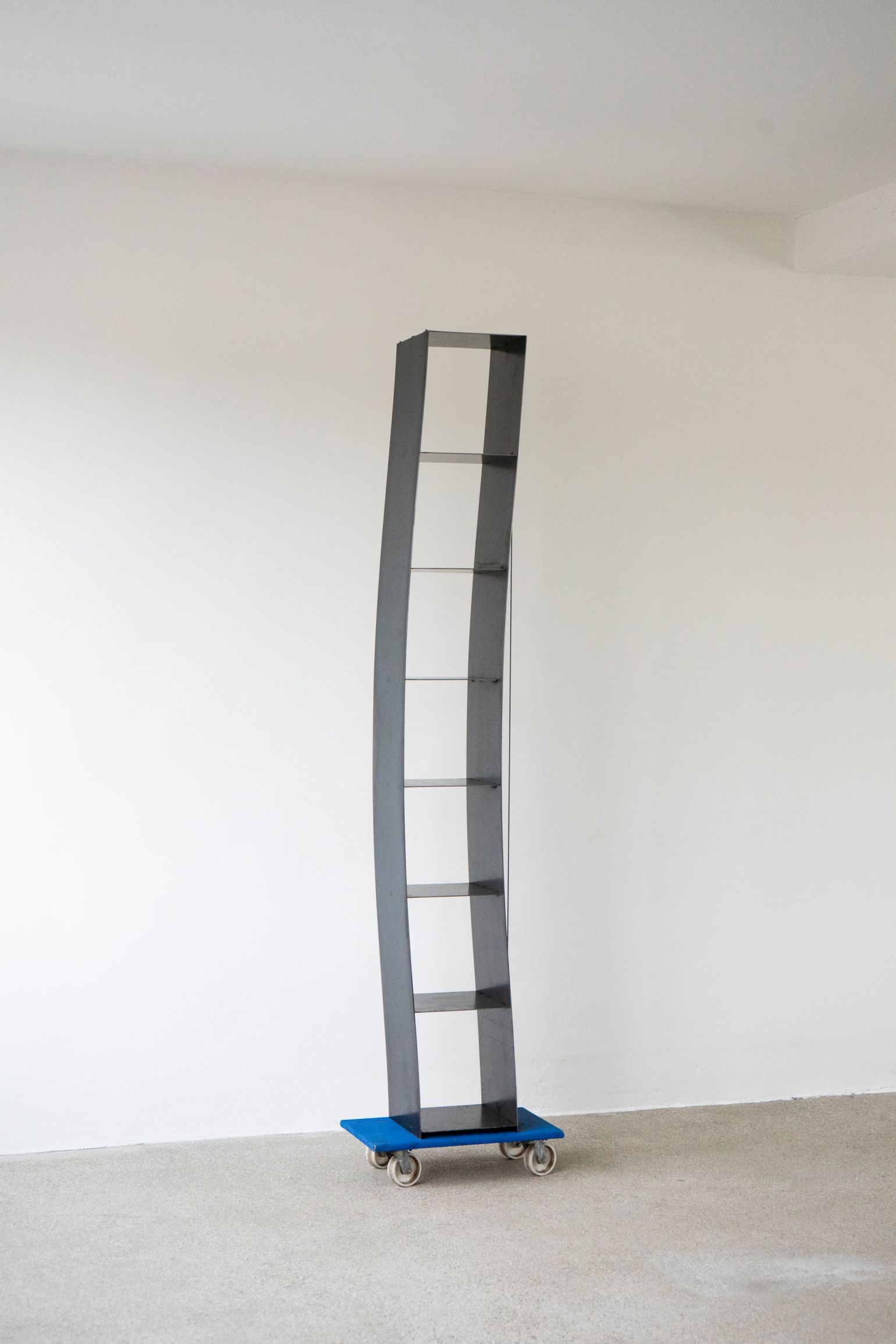 Gespanntes Regal von Moormann: Modernes, graues Bücherregal mit Glasböden auf blauem Rollbrett vor weißer Wand.