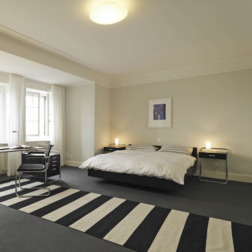 Schlafzimmer mit Thonet B 117 Beistelltischen mit Schublade neben einem Bett und Schreibtisch.