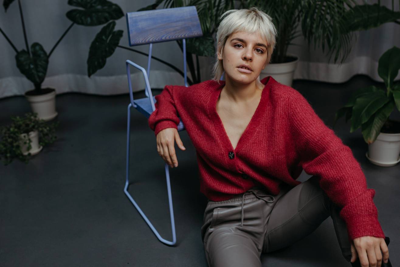 Frau lehnt an blauem Stuhl mit Armlehne. Roter Cardigan und graue Hose. Zimmerpflanzen im Hintergrund.