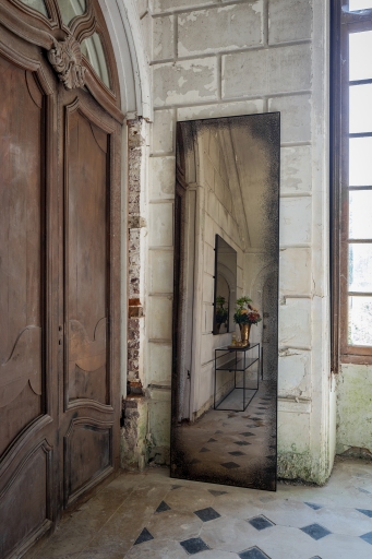 Ethnicraft Wandspiegel "heavy aged bronze": Rechteckiger Spiegel mit Antik-Finish, stehend in einem Raum mit Tür und Fenster.