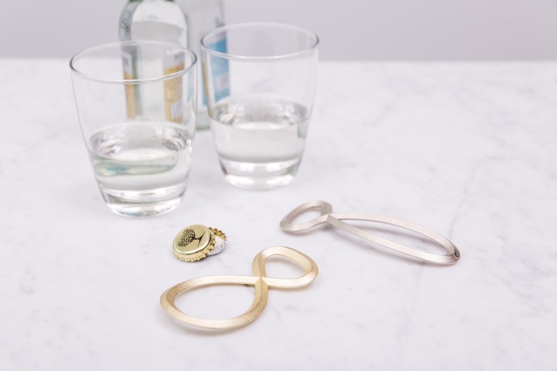 Poesie et Table Flaschenöffner Flaschenfreund silber Räder neben Gläsern und Kronkorken auf Marmor.