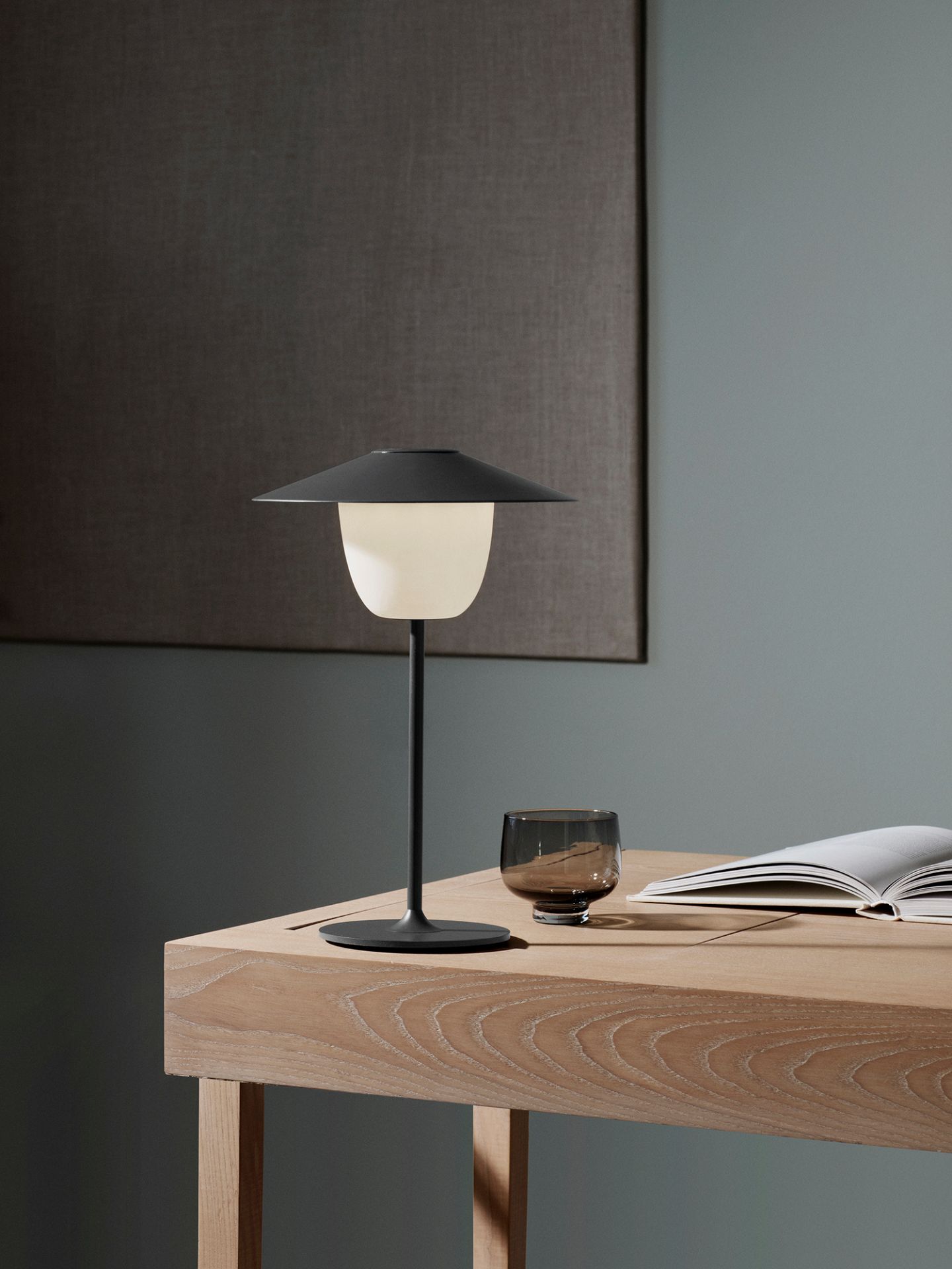 Ani Mobile LED Tischleuchte von Blomus auf einem Holztisch, neben einem Buch und Glas.