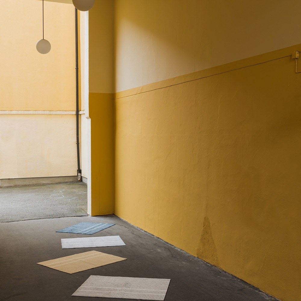 Løype Fußmatten in verschiedenen Farben für Innen und Außen, gestreiftes Design, vor gelber Wand.