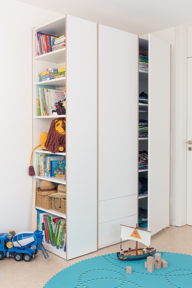 Weißes Modular Plus Regal von Müller Möbelwerkstätten im Kinderzimmer, gefüllt mit Büchern, Spielzeug und Kleidung.