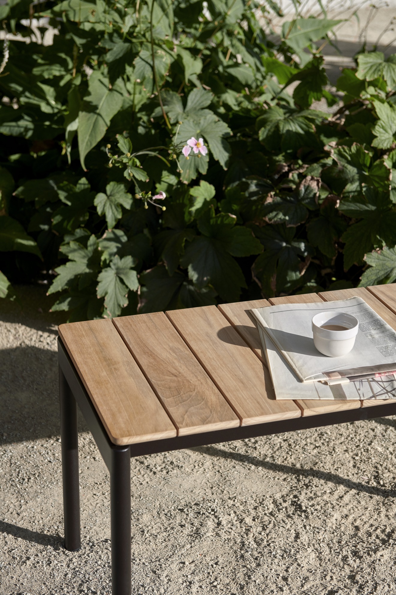 Nahaufnahme der AndTradition Ville AV27 Gartenbank aus Teakholz mit Zeitung und Kaffeetasse.