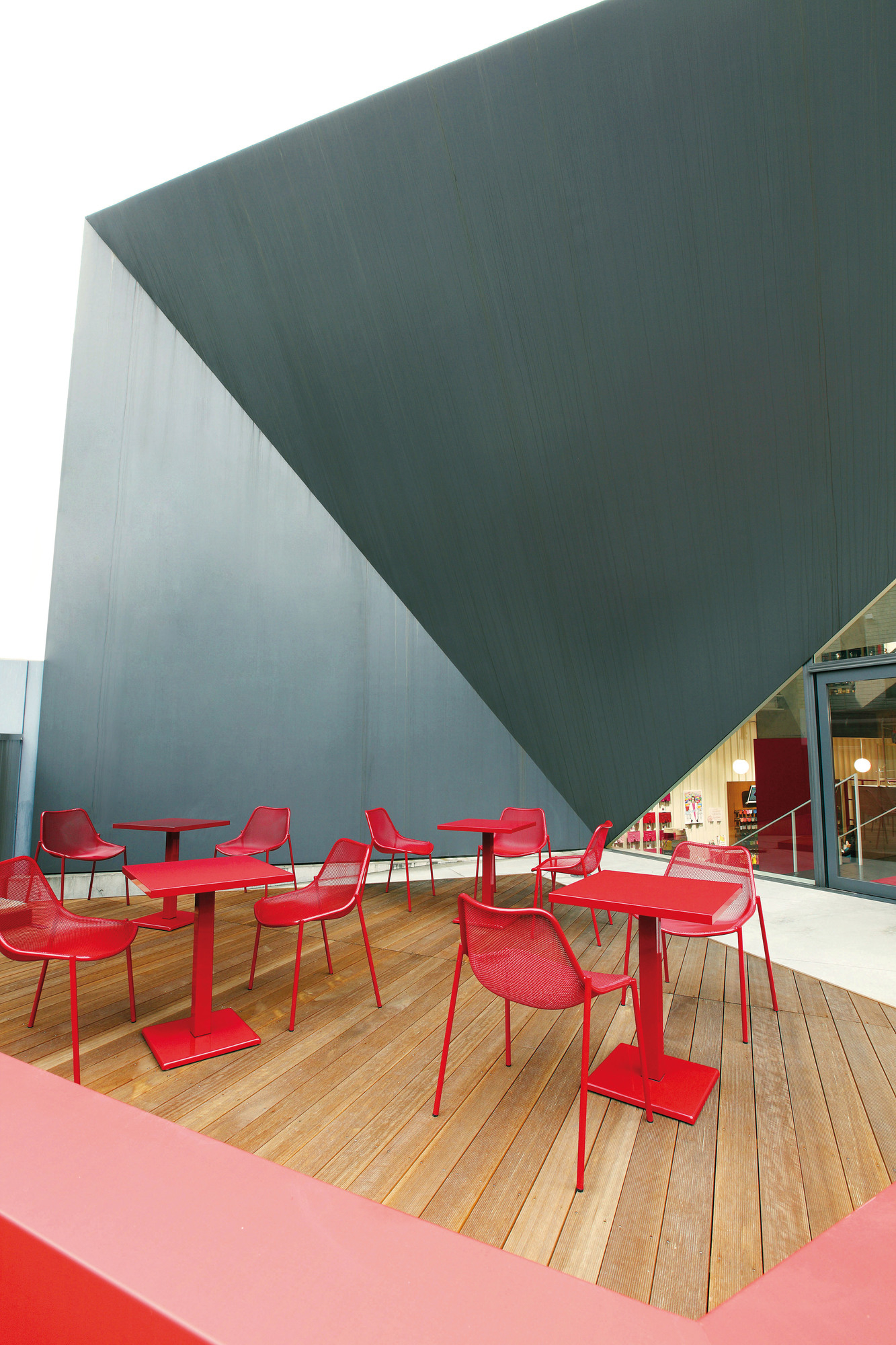 Rote Emu Stühle und Tische auf Holzterrasse vor modernem Gebäude. Outdoor Möbel.