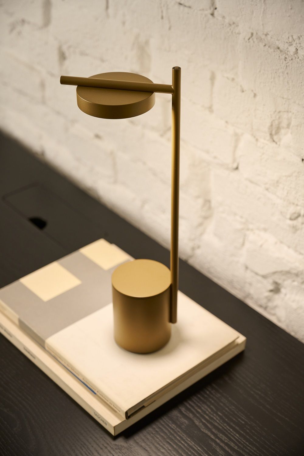 Messingfarbene Igram Akku Tischleuchte von Grupa auf Büchern vor einer Backsteinwand.