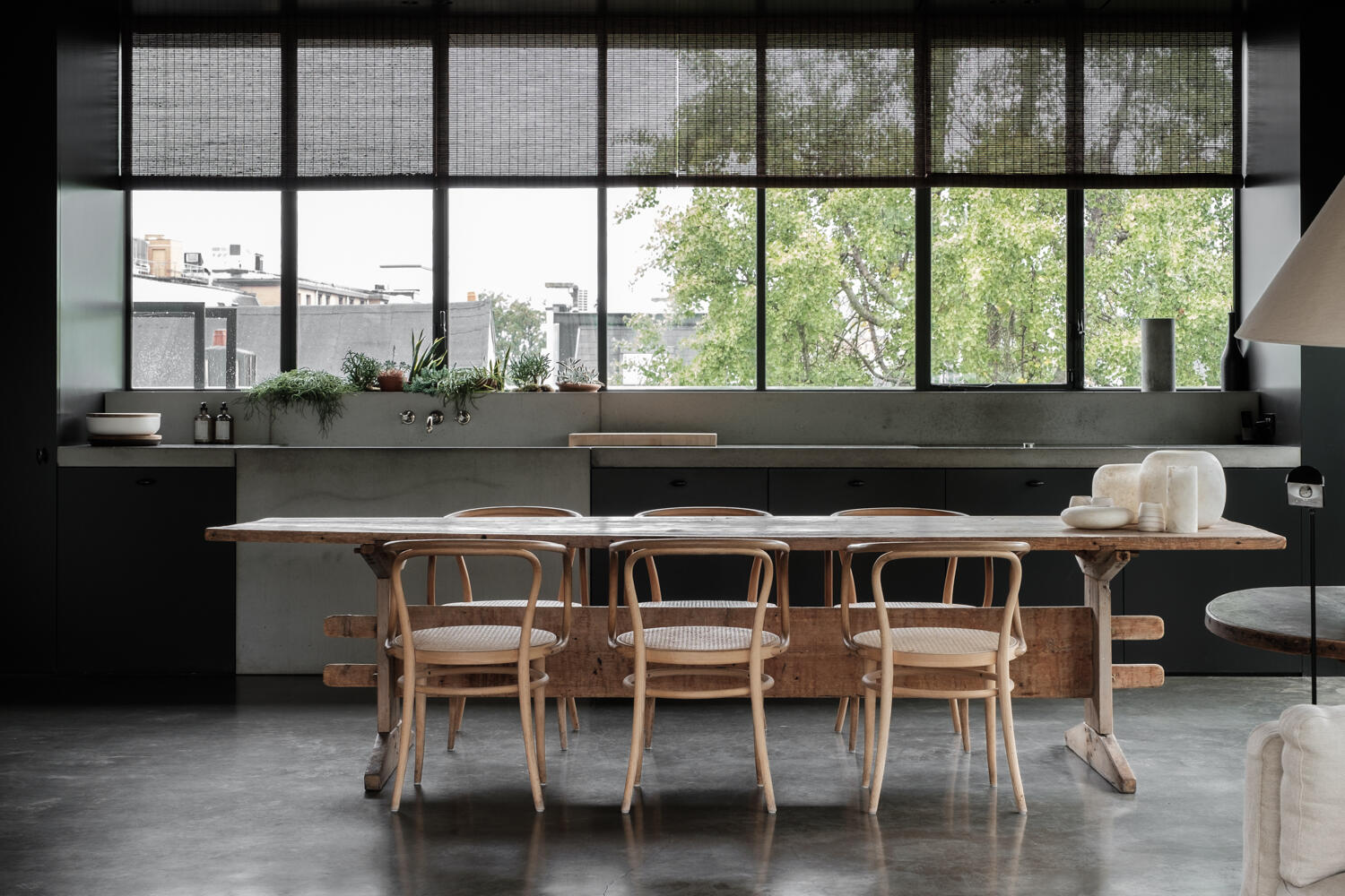 Esstisch aus Holz mit Wiener Geflecht Stühlen in modernem Loft mit Betonküche und großem Fenster.