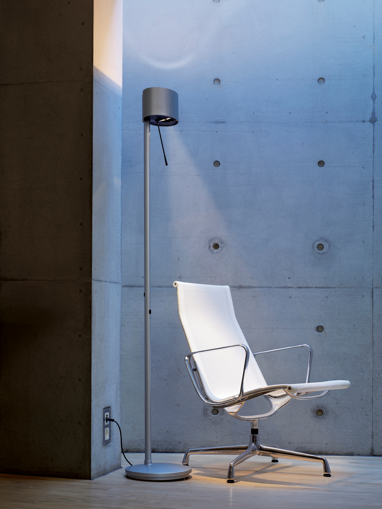 Diogenes LED Stehleuchte Belux in Goldgrau neben einem weißen Bürostuhl vor Betonwand.