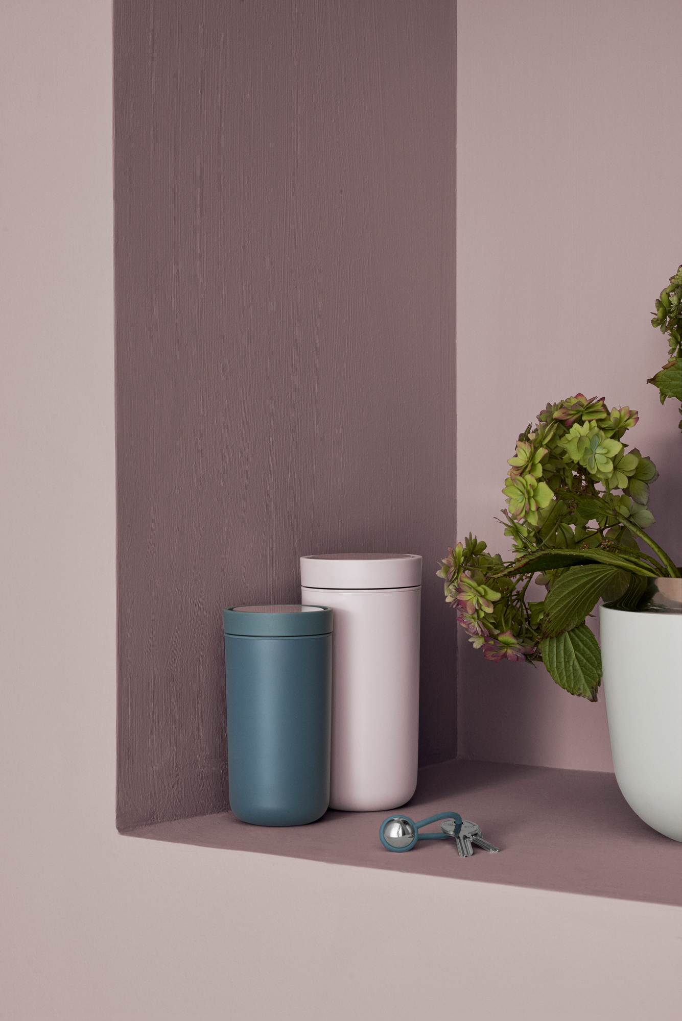 Zwei Stelton To Go Click Thermobecher in Blau und Weiß, daneben Schlüssel und Hortensie auf Regal.