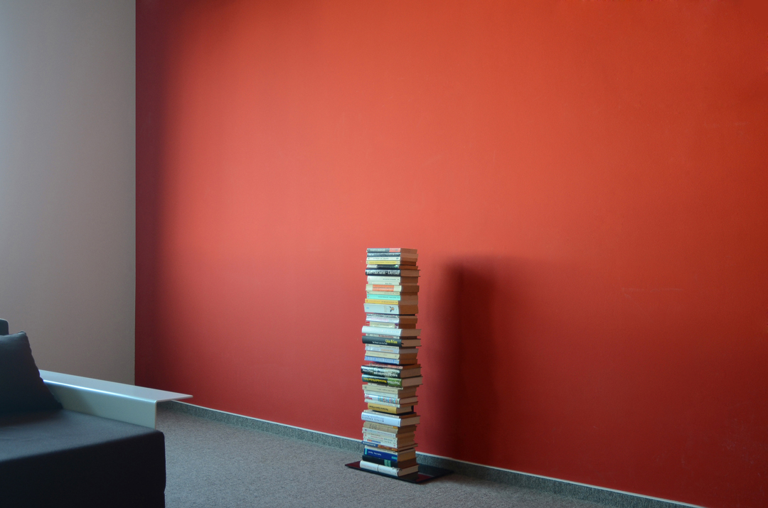 Booksbaum Standregal: Bücherstapel als Bücherregal vor roter Wand. Einzelnes, minimalistisches Design.