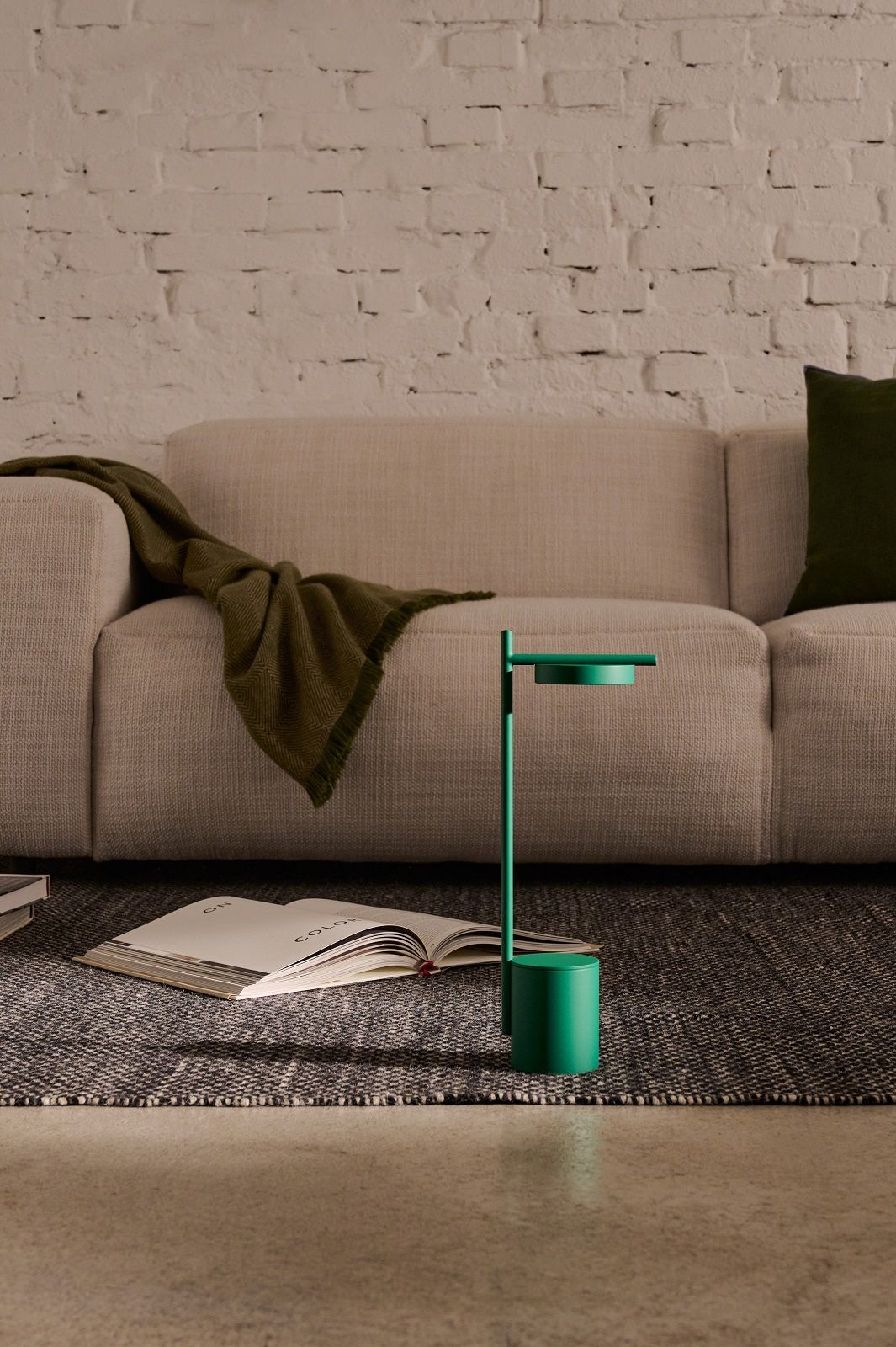 Grüne Igram Akku Tischleuchte von Grupa neben Buch auf Teppich, Sofa im Hintergrund.