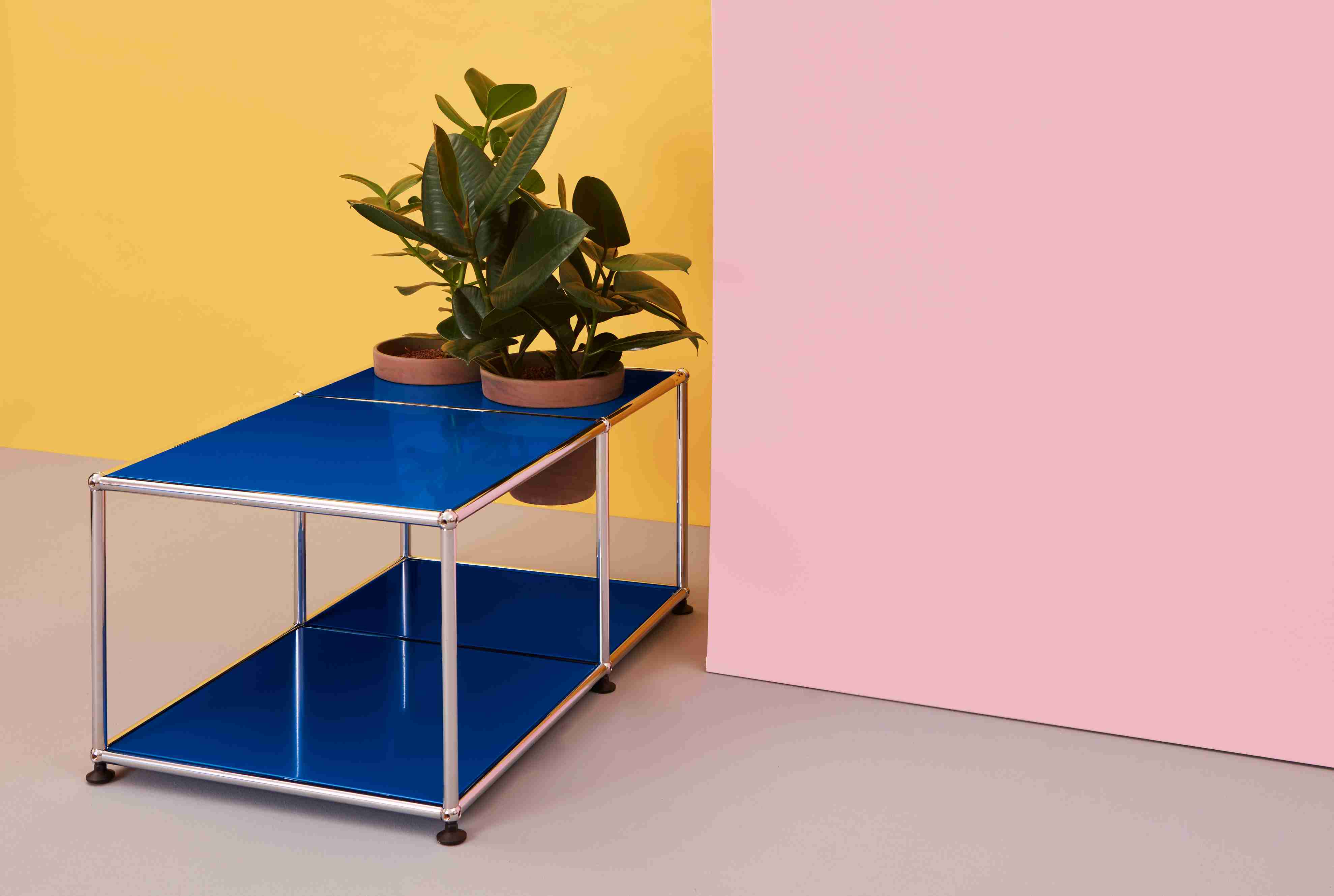 Blauer USM Haller Beistelltisch mit Pflanzen auf dem Tisch vor gelbem und rosa Hintergrund.