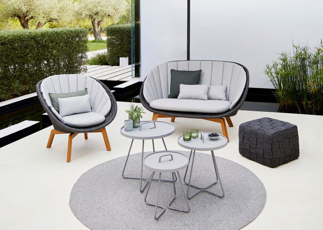 Graue Gartenmöbel-Lounge mit Sofa, Sessel, Beistelltischen und Pouf auf einem runden Teppich im Außenbereich.