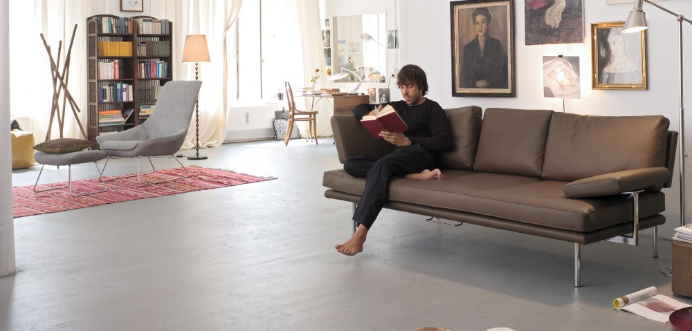 Mann liest auf braunem Ledersofa von Walter Knoll mit verstellbaren Armlehnen. Wohnzimmer-Szene.