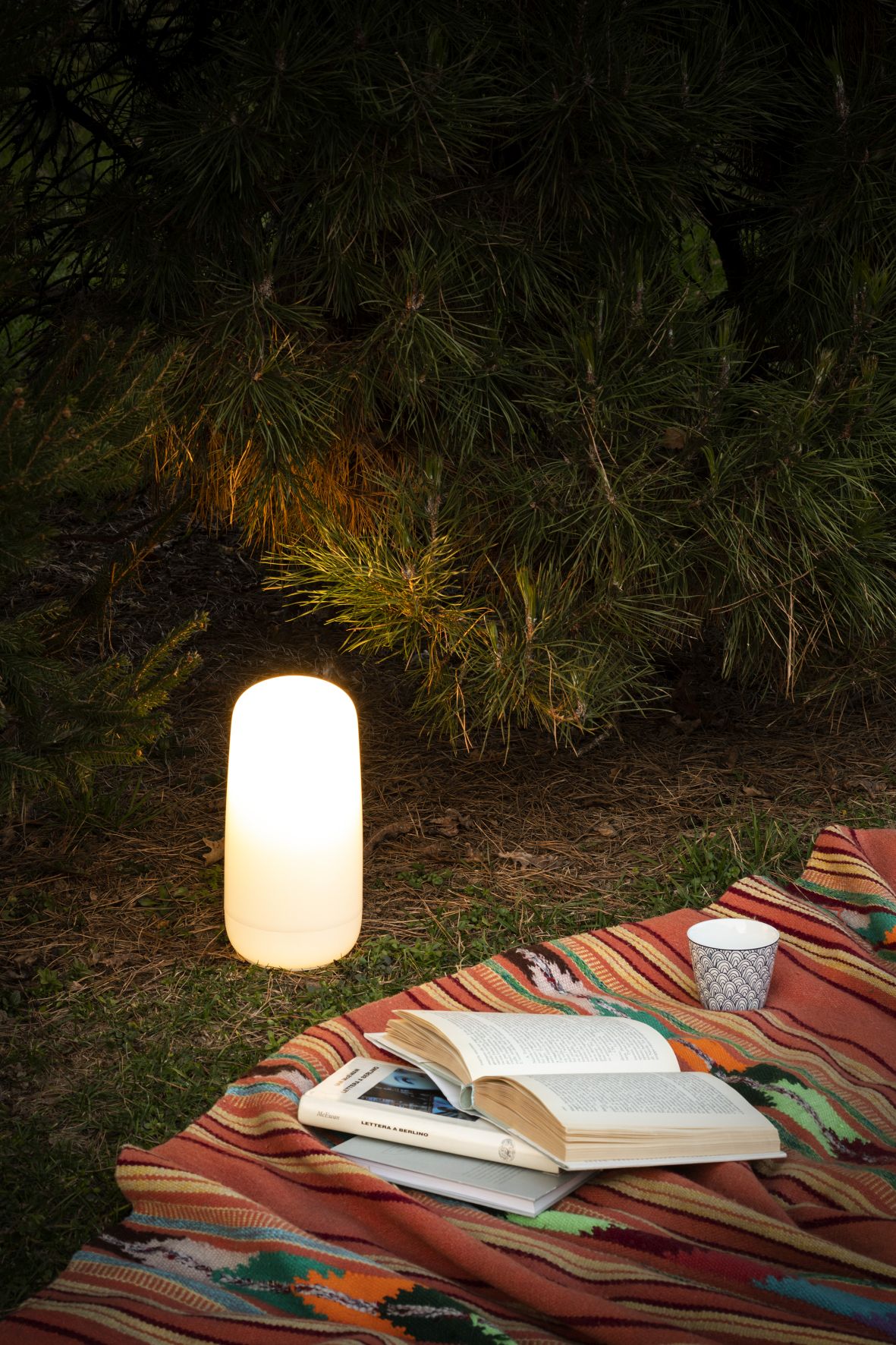 Artemide Gople Portable Akkuleuchte auf einer Decke im Freien, neben Büchern und einer Tasse.
