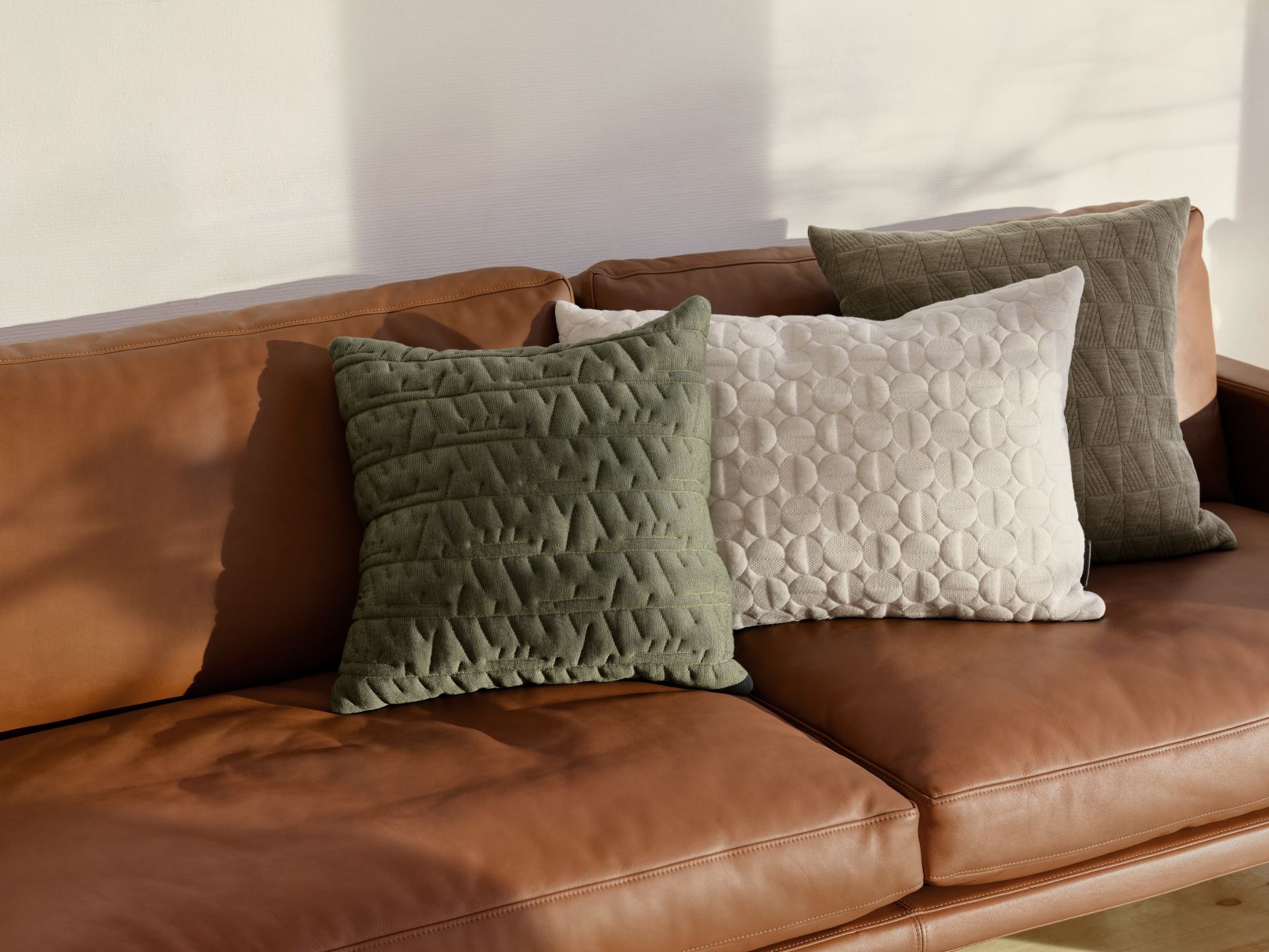 Nahaufnahme von drei dekorativen Kissen auf einem Ledersofa, darunter das grüne Tassel AJ Cushion Kissen von Fritz Hansen.