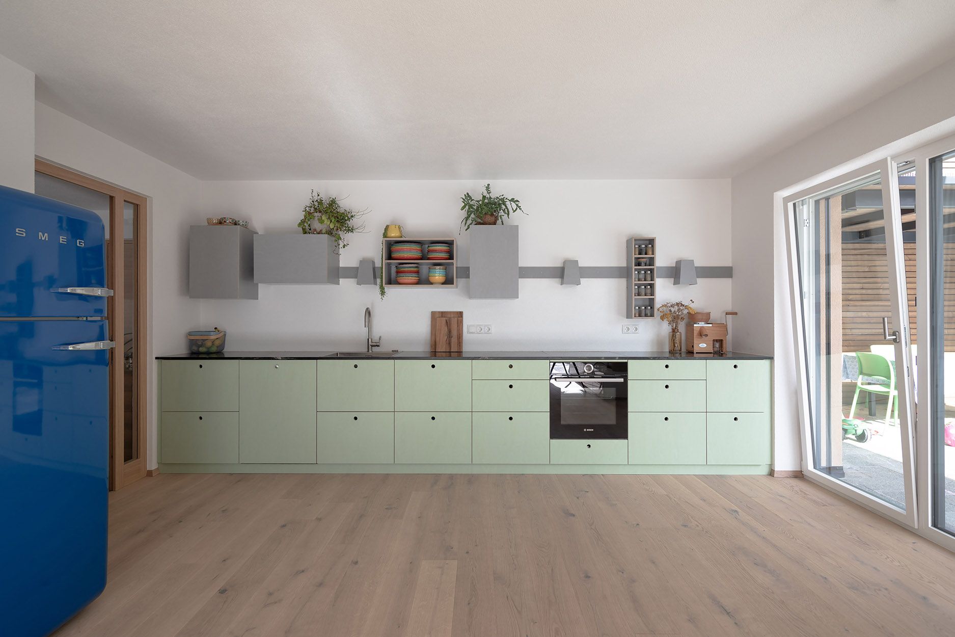 Moderne Küche mit pastellgrünen Schränken, schwarzer Arbeitsplatte, blauem SMEG Kühlschrank und Holzboden.