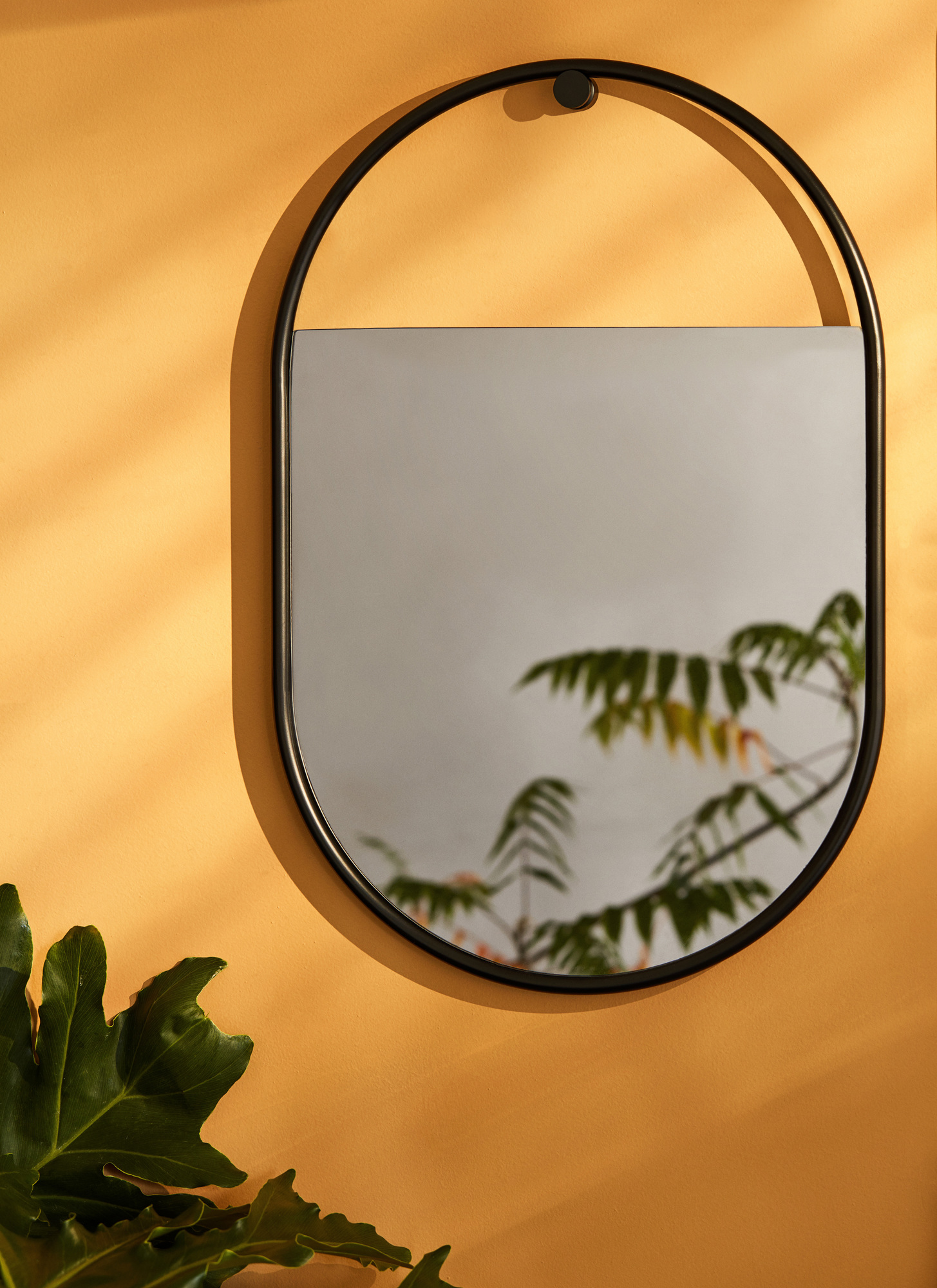 Ovaler Peek Wandspiegel mit schwarzem Rahmen an ockerfarbener Wand, spiegelt Baum. Modernes Design.