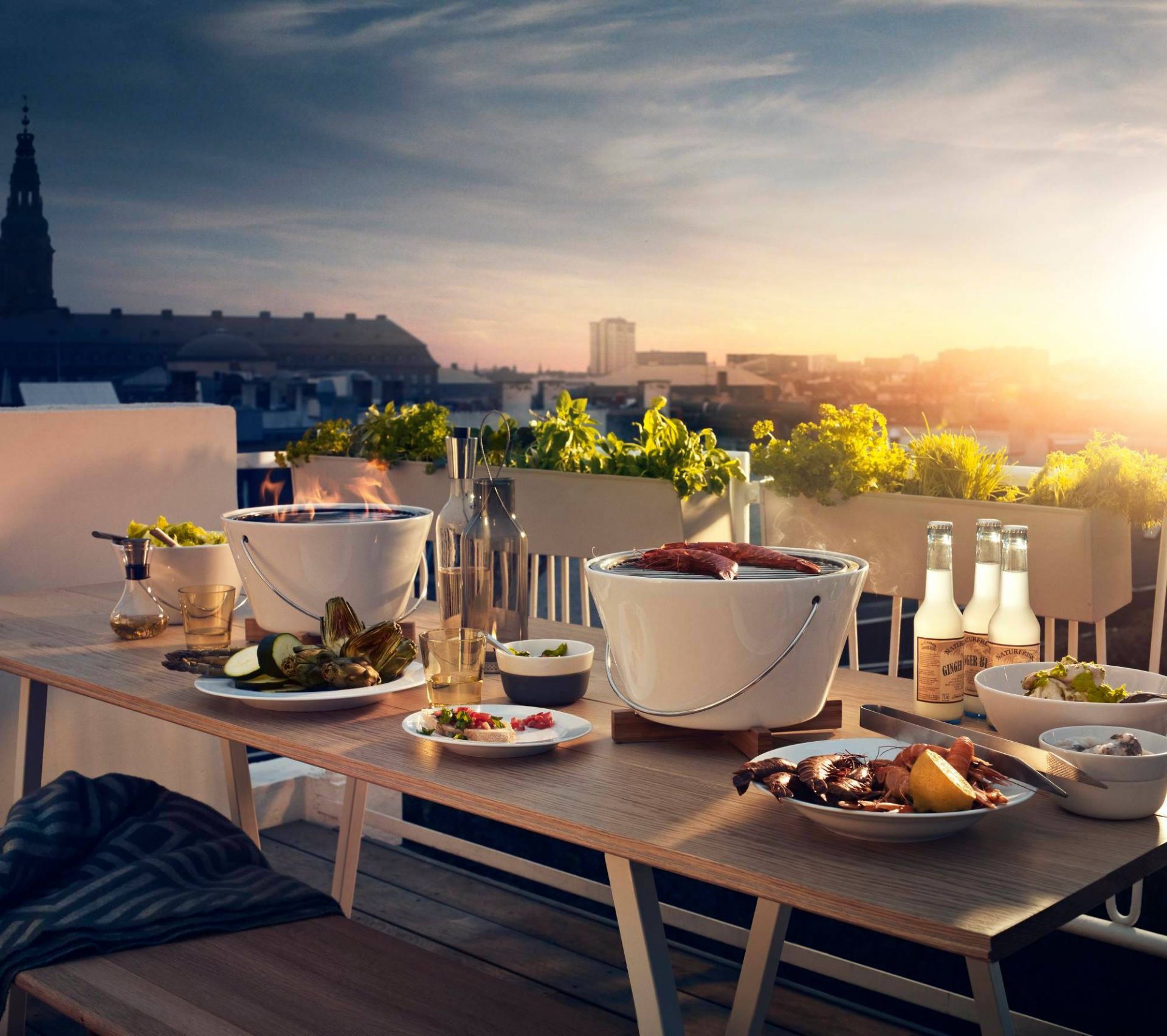 Grillabend auf der Dachterrasse: Zwei weiße Holzkohlegrills mit Speisen, Getränken und Blick auf die Stadt.