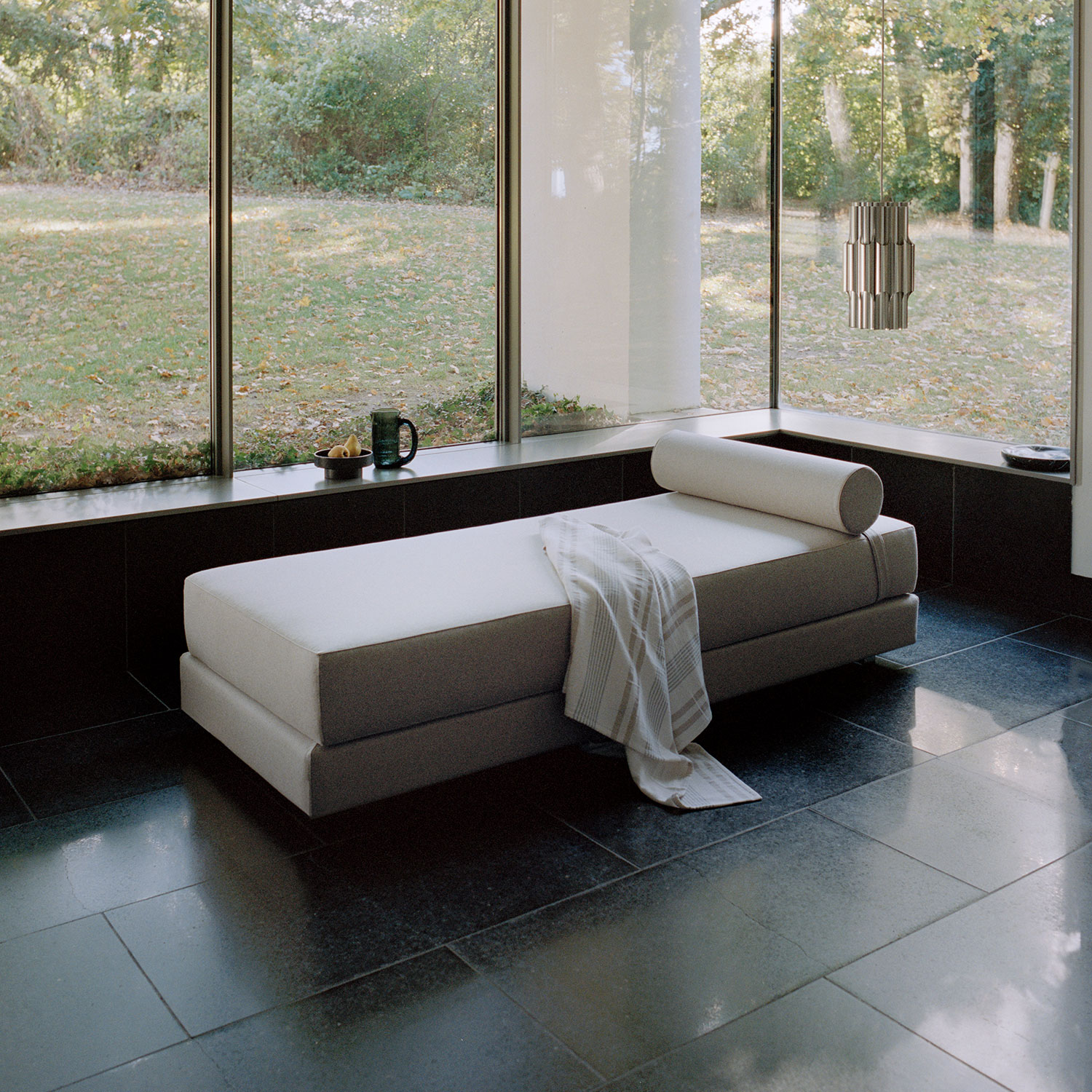 Helle moderne Liege mit Kissen und Decke vor großem Fenster mit Gartenblick.