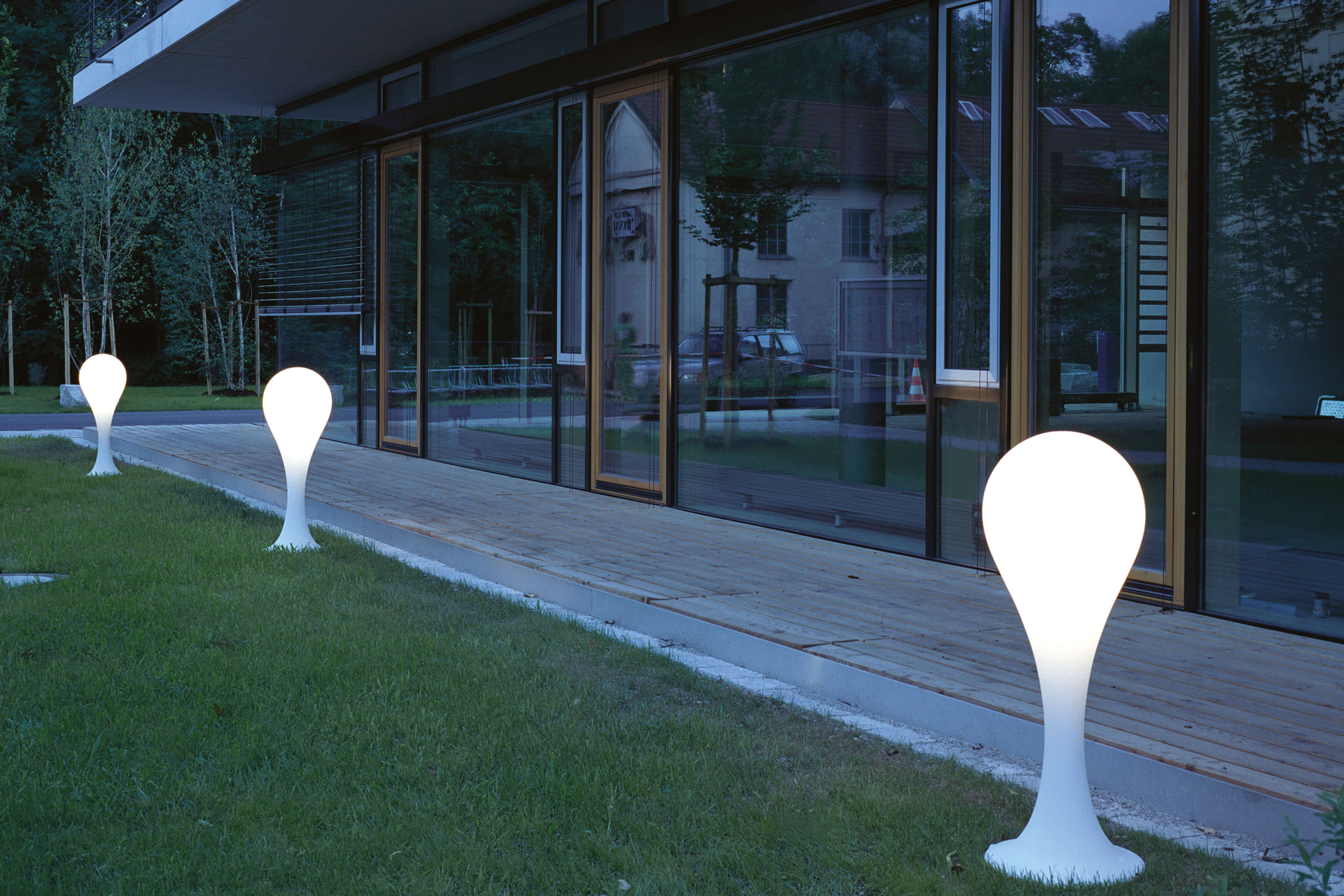 Drei Liquid Light Drop Outdoor Standleuchten von Next auf Rasen vor einem Gebäude.