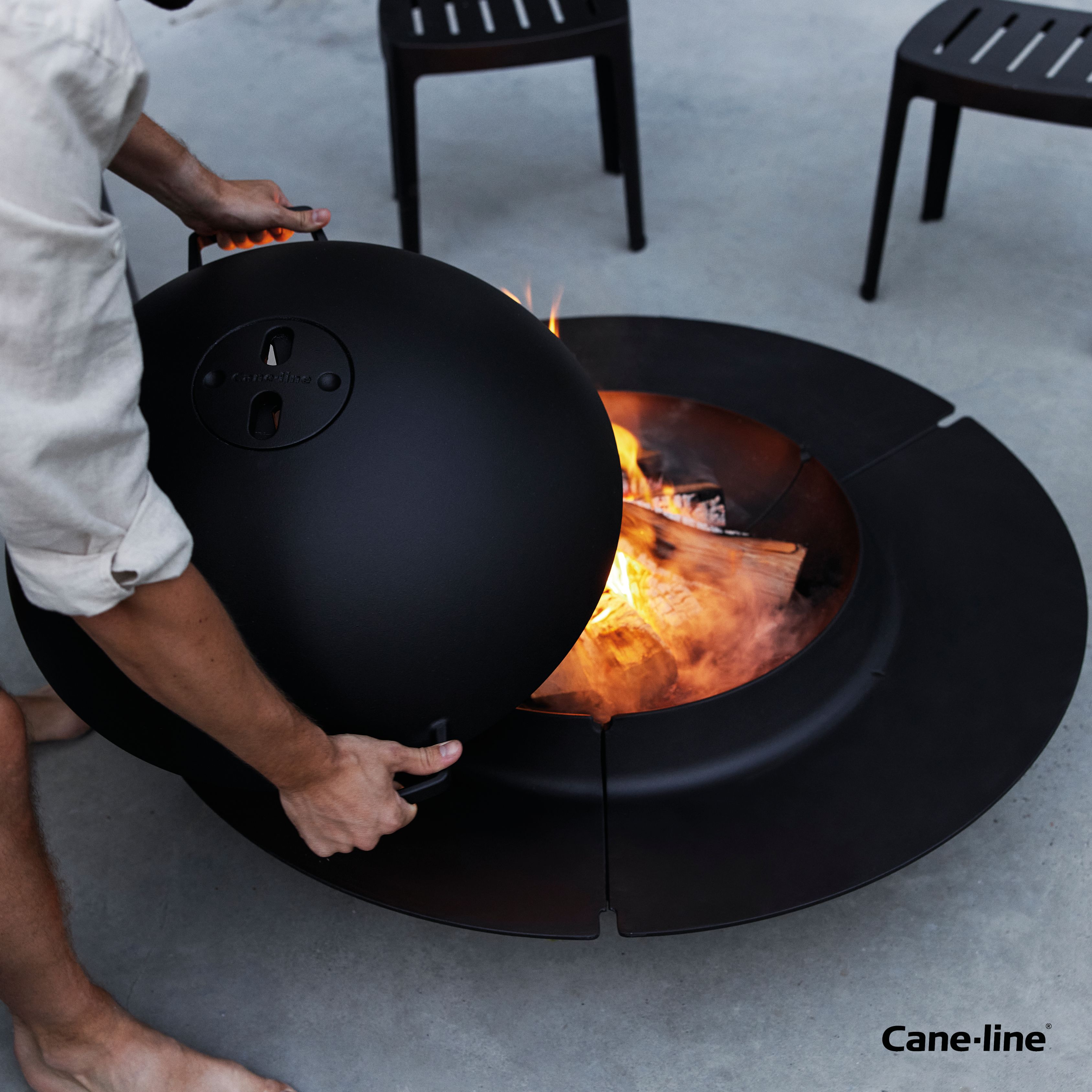 Schwarze Ember Feuerschale von Cane-Line mit Deckel wird geöffnet, Feuerholz und Flammen sichtbar.