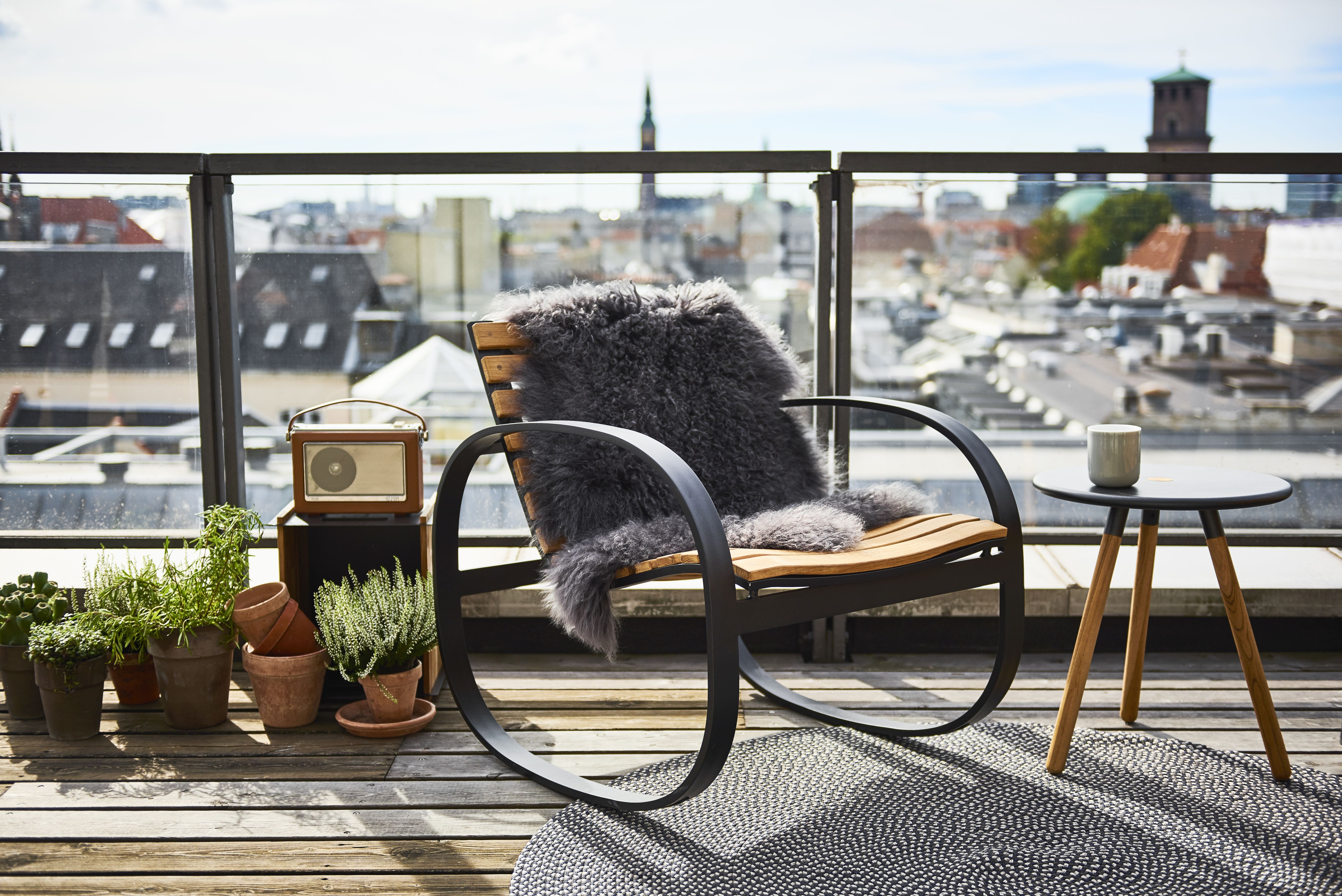Parc Schaukelstuhl von Cane-Line auf einer Dachterrasse mit Schaffell und Beistelltisch. Outdoor Möbel für entspannte Momente.