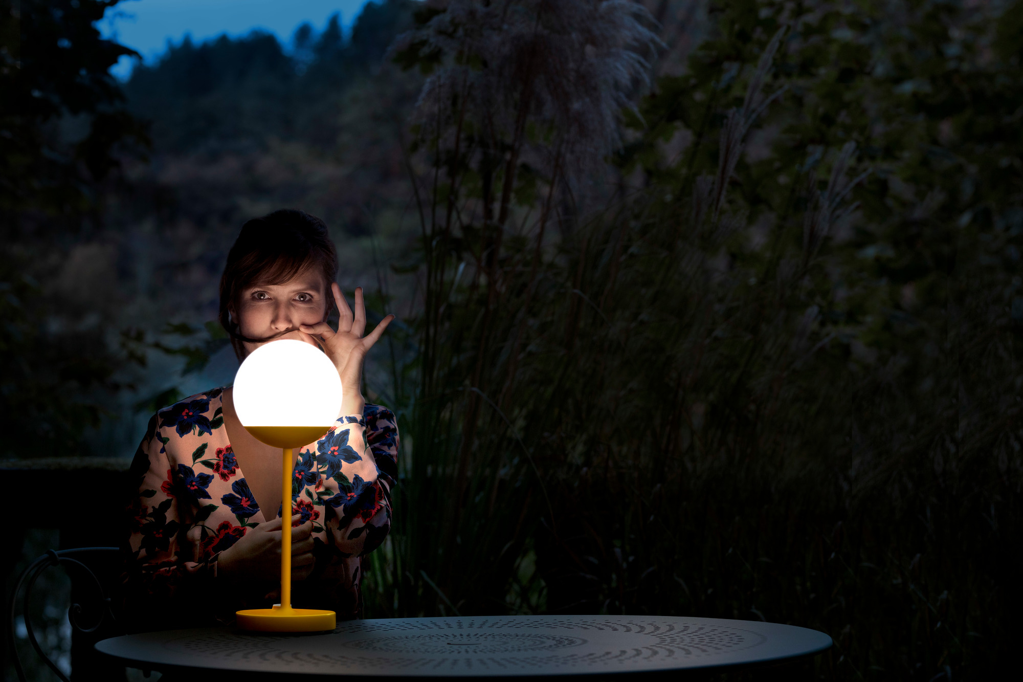 Mooon Tischleuchte von Fermob, anthrazitfarbener Tisch, Frau mit Blumenmuster-Bluse im Garten. Dekorative Leuchte für stimmungsvolle Beleuchtung.