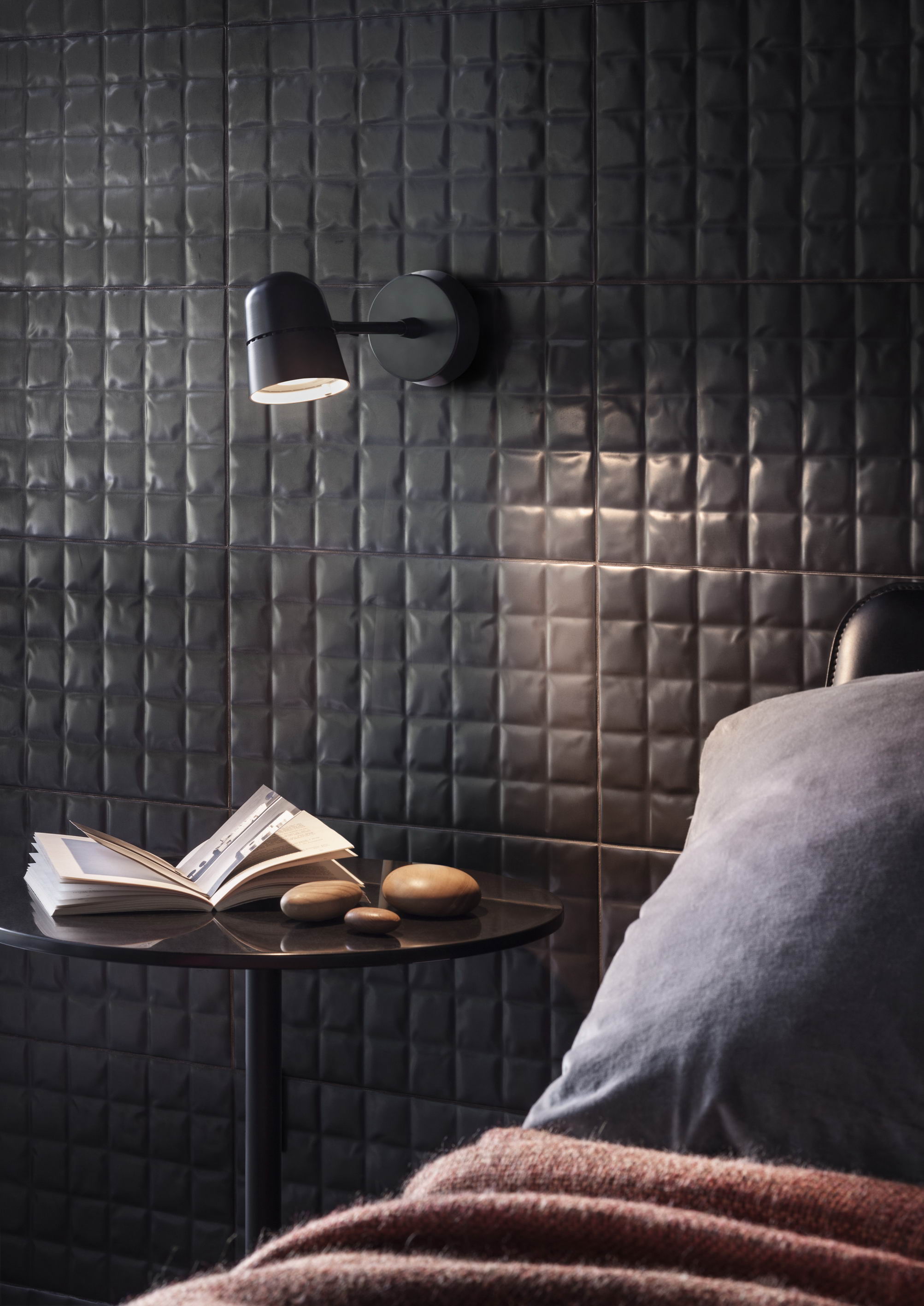 Schwarze Luceplan Spot Wandleuchte neben Bett mit Buch und Deko-Steinen auf Nachttisch.