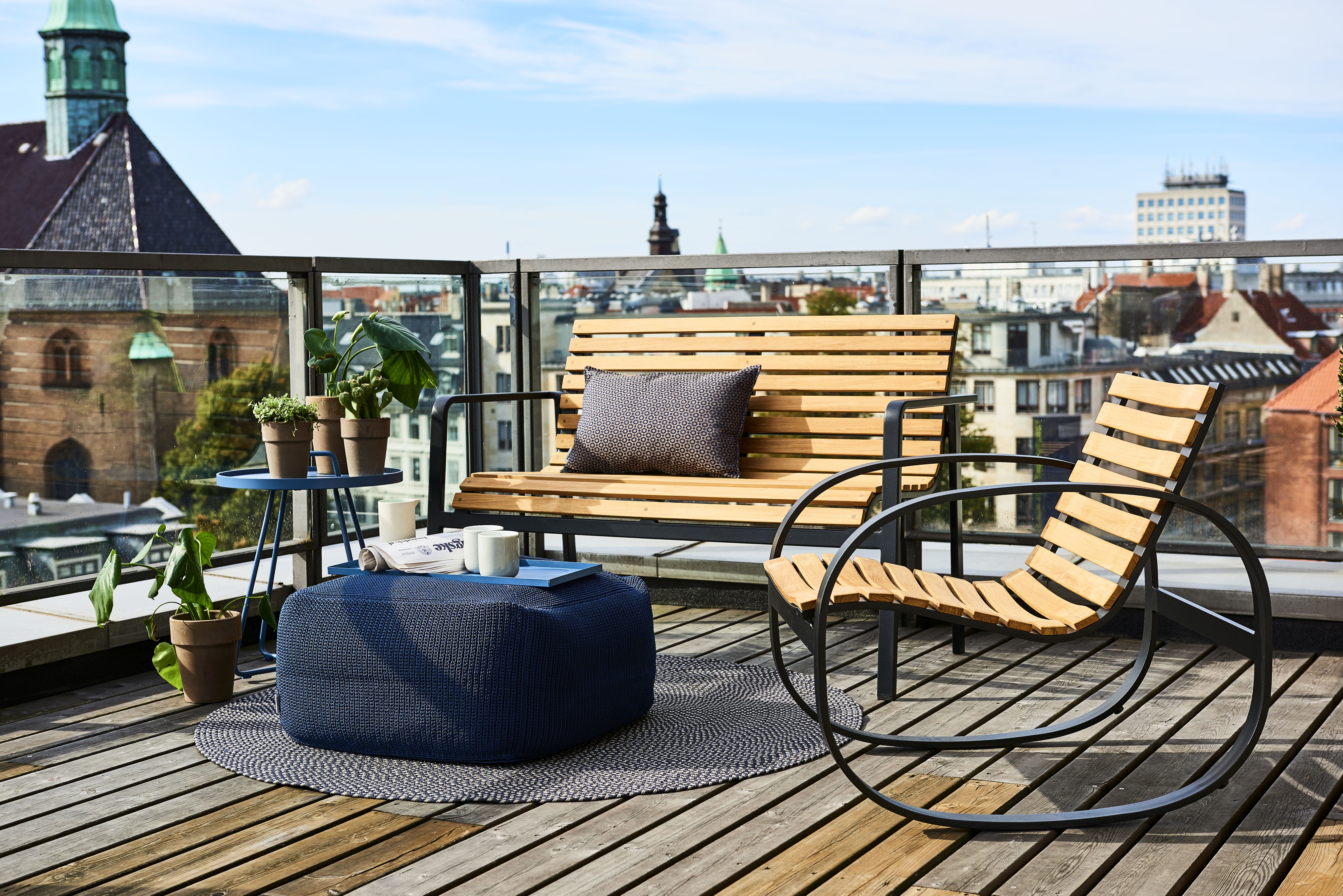 Parc Schaukelstuhl von Cane-Line aus Teakholz und Metall auf einer Dachterrasse mit Stadtblick.